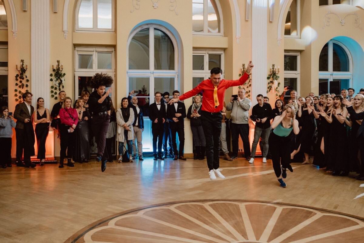 W dużej, eleganckiej sali gromadzi się grupa ludzi, aby obejrzeć energetyczny występ studniówki. Tancerze podskakują w powietrze, na co formalnie ubrana publiczność patrzy z entuzjazmem i owacjami, tworząc żywy reportaż ze studniówki.