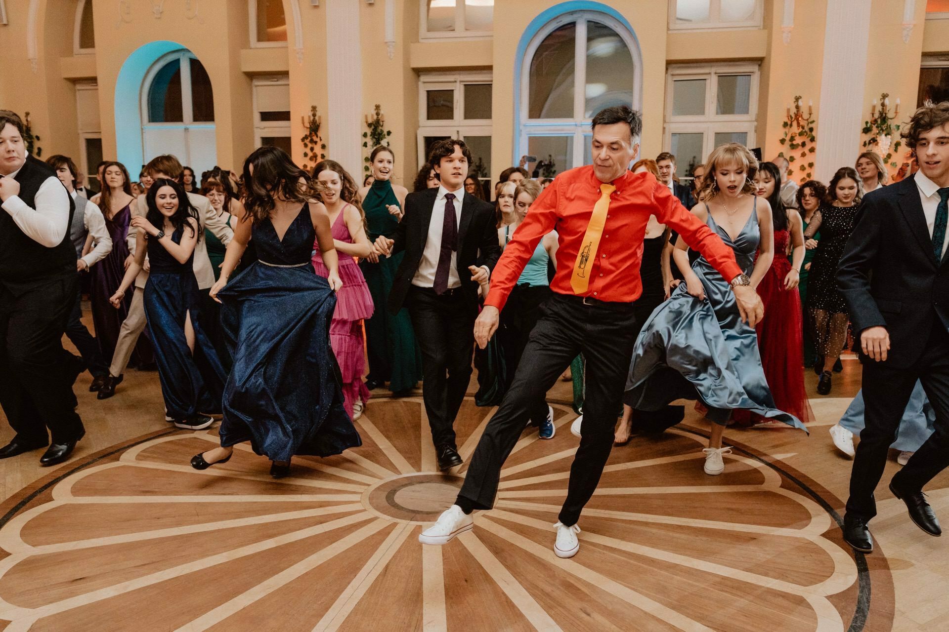 A group of people in formal attire are dancing joyfully in a spacious, elegant room with arched windows and warm lighting. The man in the middle is wearing a red shirt and yellow tie and energetically leads the dance. Others follow in this colorful "prom rep" with big smiles, lifting gowns and suits.  