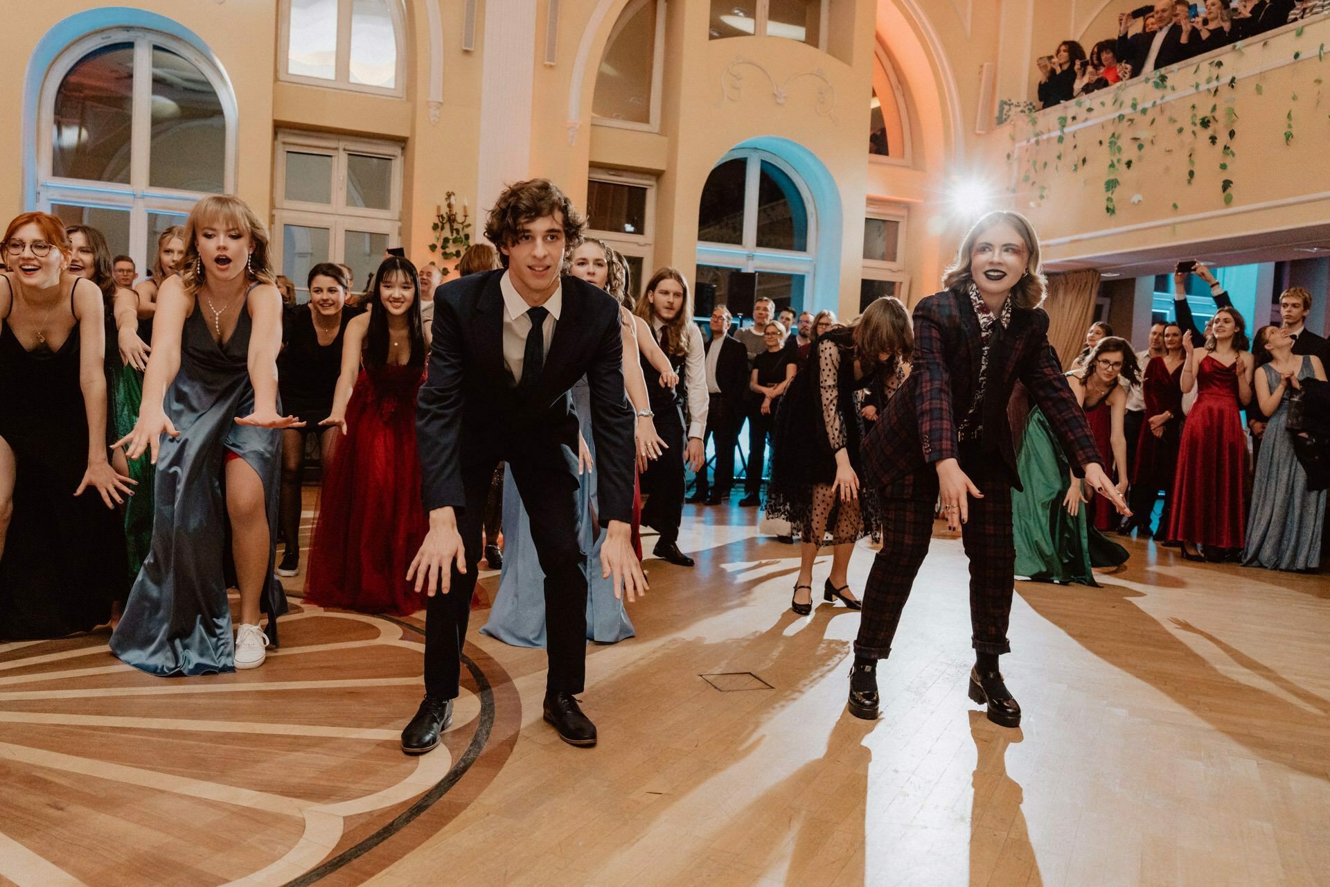 A group of people dressed in formal attire are dancing in a large, elegant room with arched windows and high ceilings. They are in synchronized formation, with arms outstretched and faces expressing enthusiasm. The prom report captures the spectators gathered around watching the dance.  