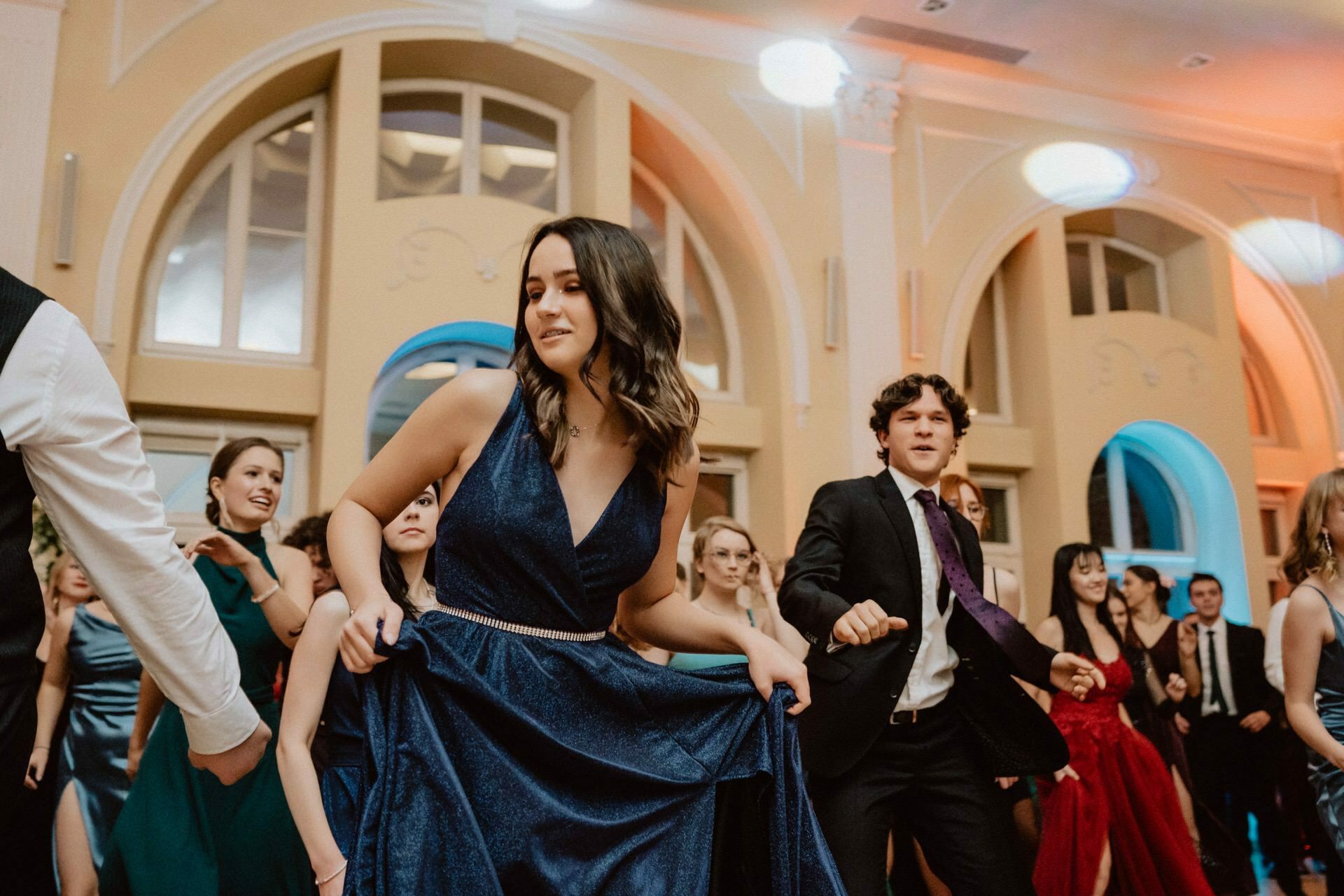 A woman in a blue dress and a man in a suit dance at a formal event, while in the background a crowd of people in elegant attire. The venue features high arches and warm lighting. The atmosphere seems lively and festive, reminiscent of a prom reportage.  