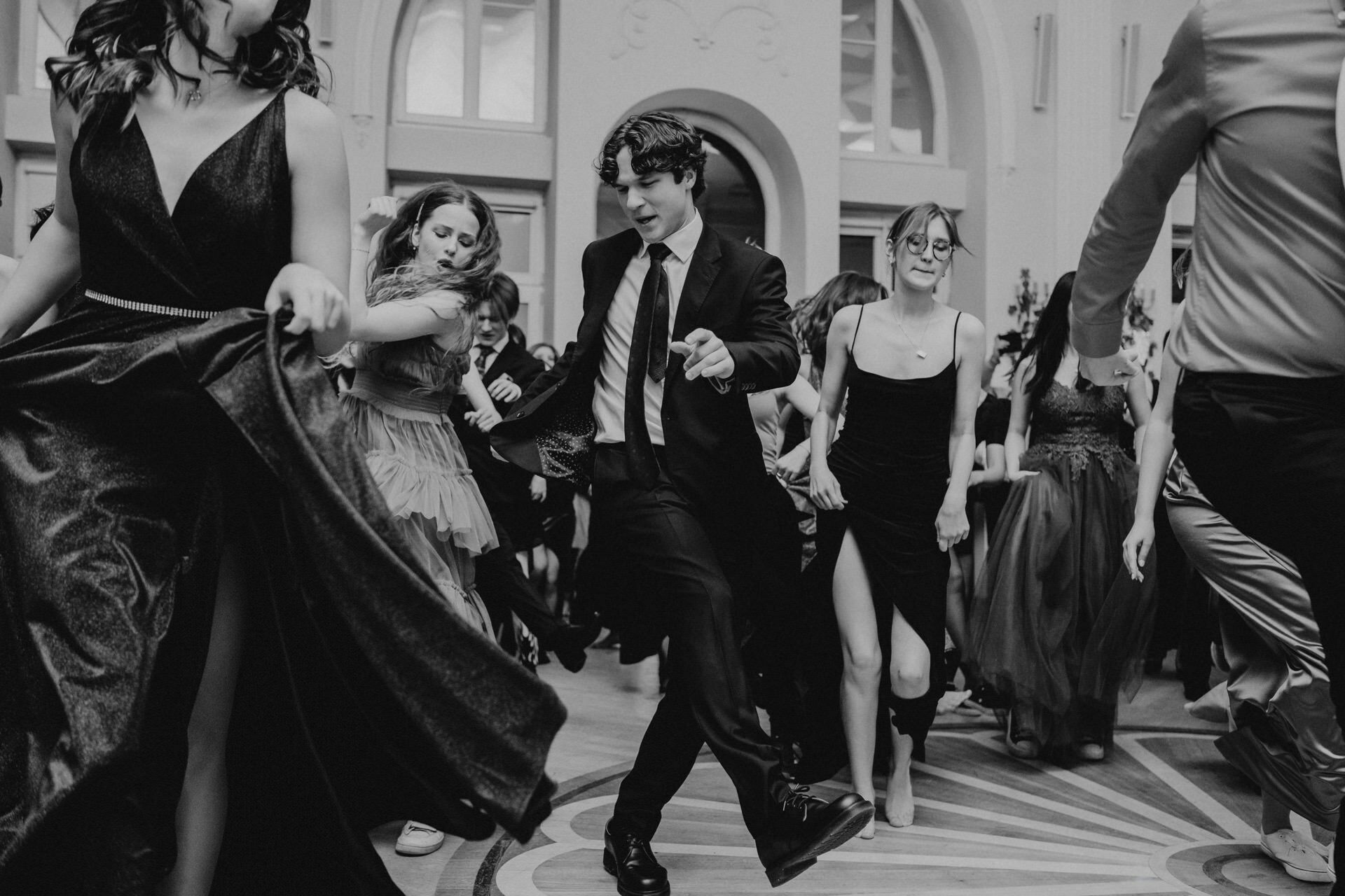 A black and white photo of a bustling dance floor at a formal event. Men and women in elegant attire, including dresses and suits, joyfully dance together. In the background, large arched windows add to the grand atmosphere - reminiscent of a prom reportage.  