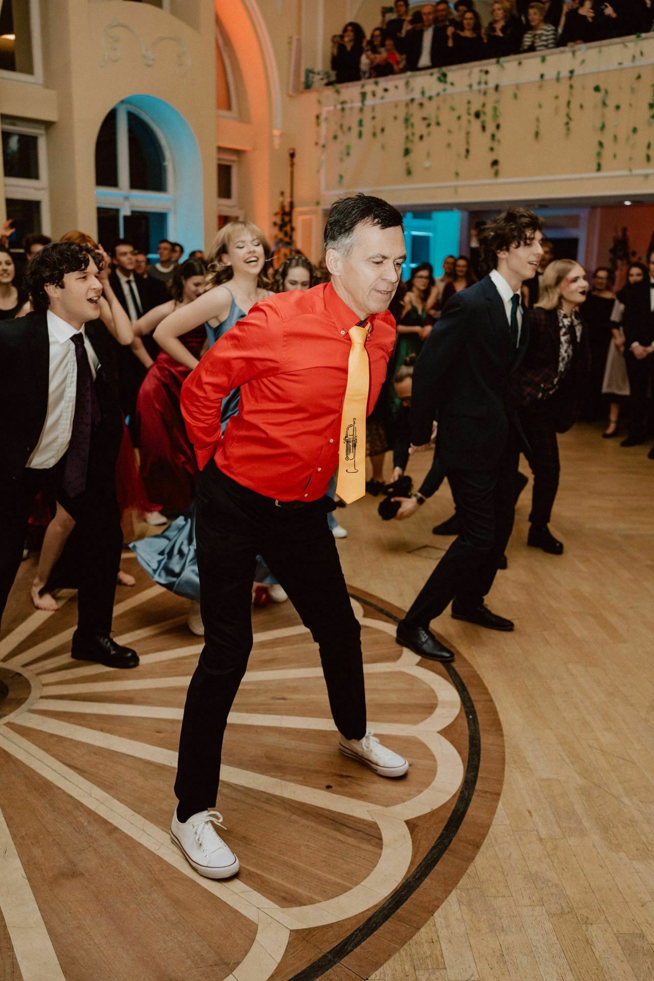 A group of people in formal attire are dancing together at a party held at an elegant venue. A man in a bright red shirt and yellow tie stands in the middle, smiling. The crowd behind them watches and enjoys the performance on the semicircular wooden floor, capturing a moment of true prom reportage.  