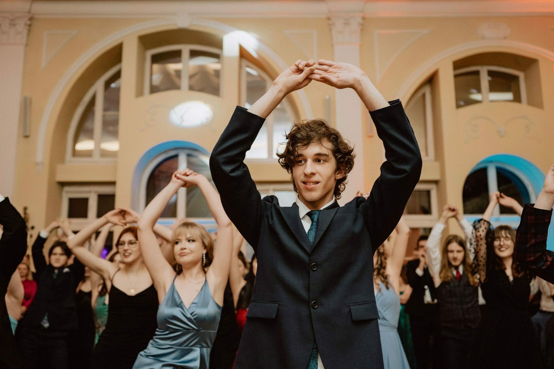 A young man in a suit dances energetically with his hands raised above his head, leading a large group of people behind him, all imitating his movements. The backdrop of the vibrant reportage of the prom is a stylish, well-lit hall with arched windows. 