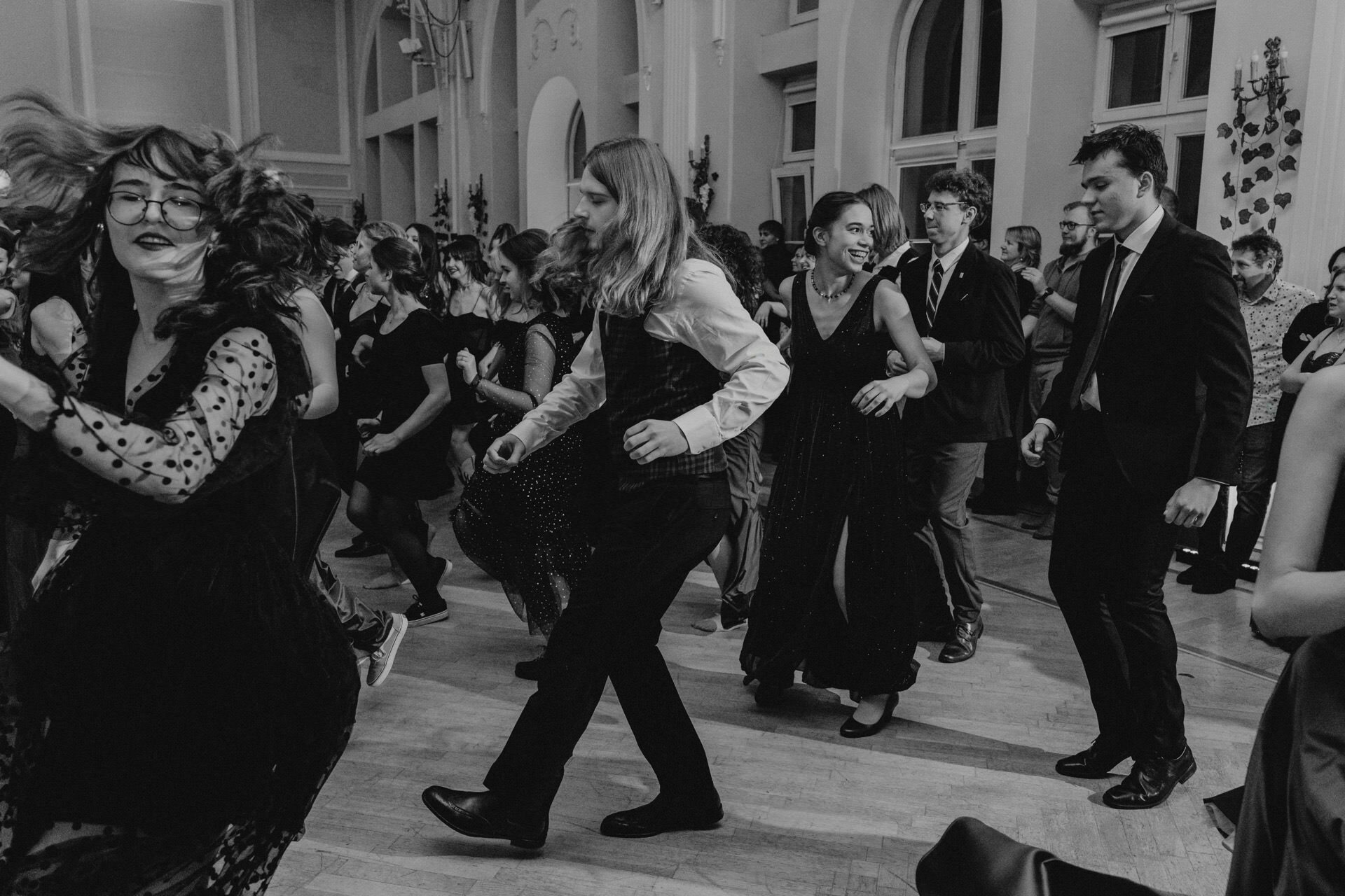 A lively group of people dance energetically in a large, elegant hall, all dressed in formal attire. The black-and-white photo is reminiscent of *a prom reportage*, capturing the dynamic movement of the dancers, the airy outfits and animated facial expressions, giving a sense of joy and celebration. 