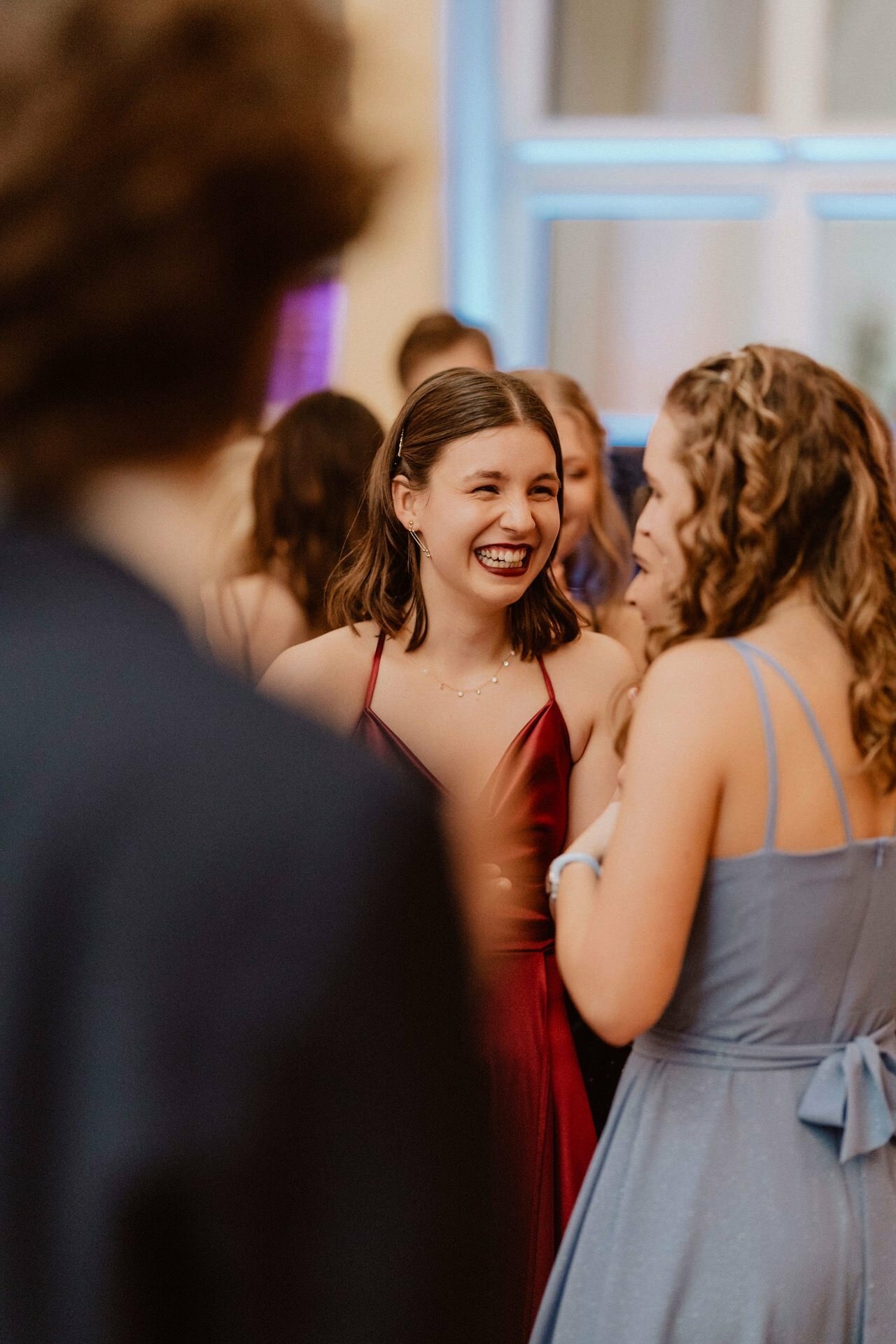 A young woman in a red sleeveless dress smiles and talks with another woman in a light blue dress at a social gathering reminiscent of a prom report. They are surrounded by other people, and a fuzzy man can be seen in the foreground, indicating that this is probably a formal or official meeting. 