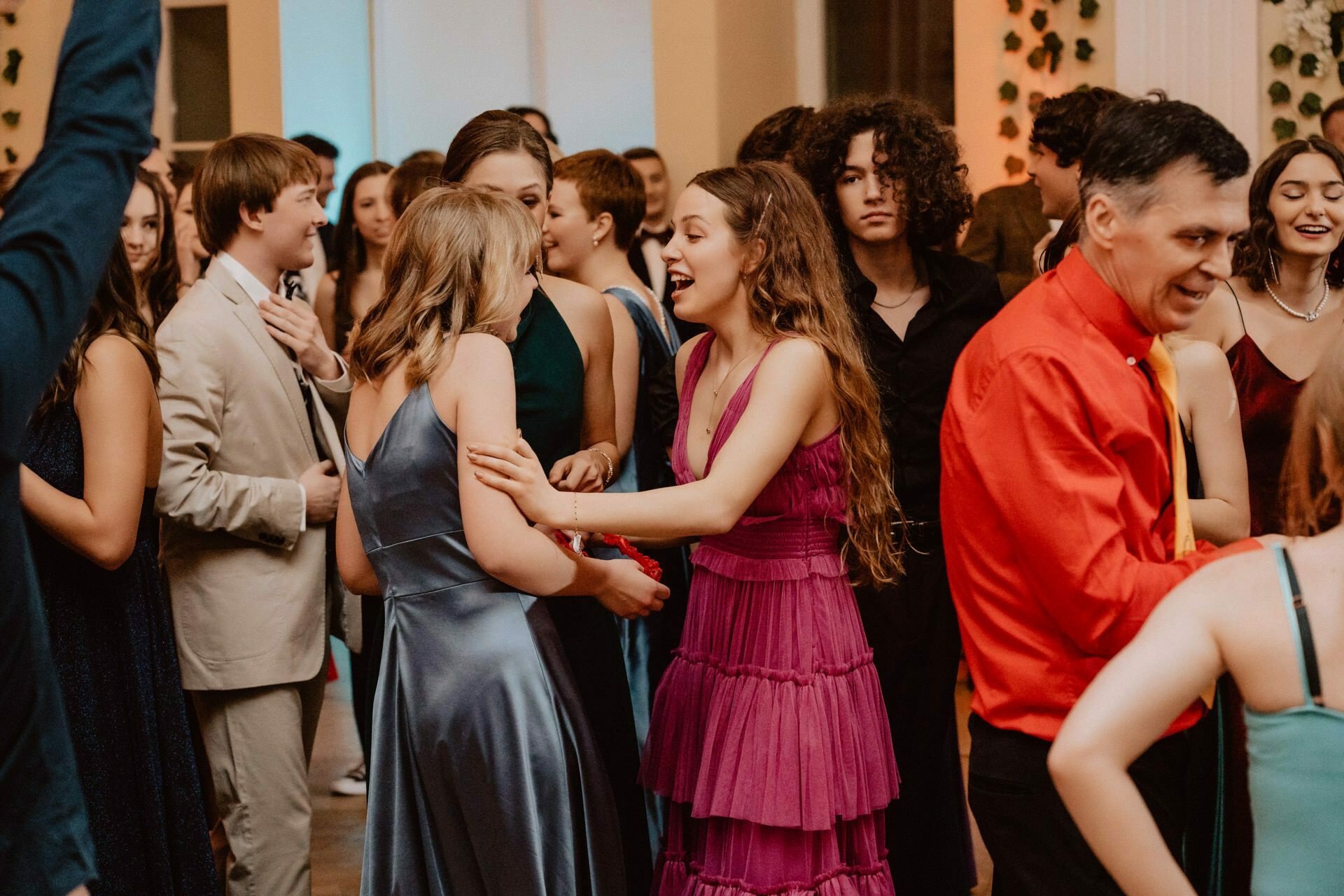 A lively group of people gather for an event held indoors, perhaps at a party or dance. They are dressed in formal attire. Two women in the foreground are engaged in a lively conversation, one in a pink dress, the other in a blue dress. This scene could be straight out of "Prom Report.   