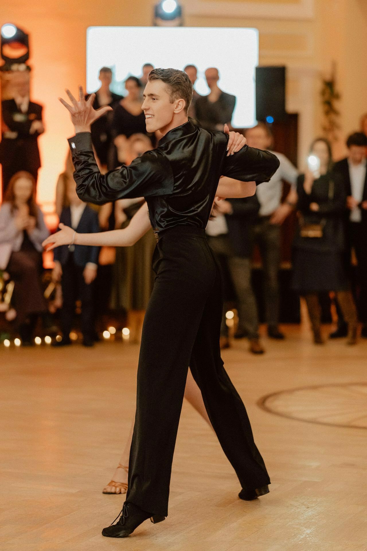 A dancer in black attire performs a dance routine with a partner who is partially visible. The man takes a dramatic pose with his hand raised and fingers extended. They are on a wooden floor and the audience watches them in the background, which resembles a reportage from a prom.  