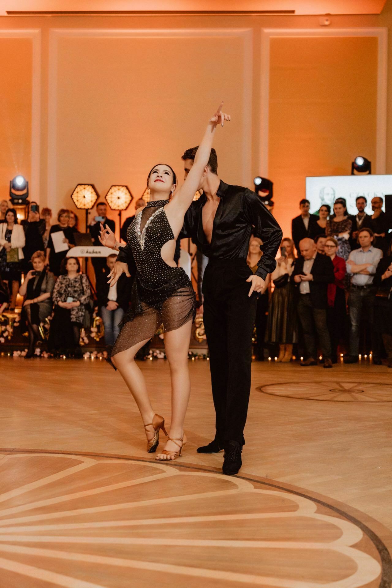 A pair of dancers perform a passionate ballroom dance on a wooden floor, reminiscent of a prom reportage, with the crowd watching in the background. The dancer adopts a dramatic pose with one arm extended upward, while the dancer stands confidently behind her. 