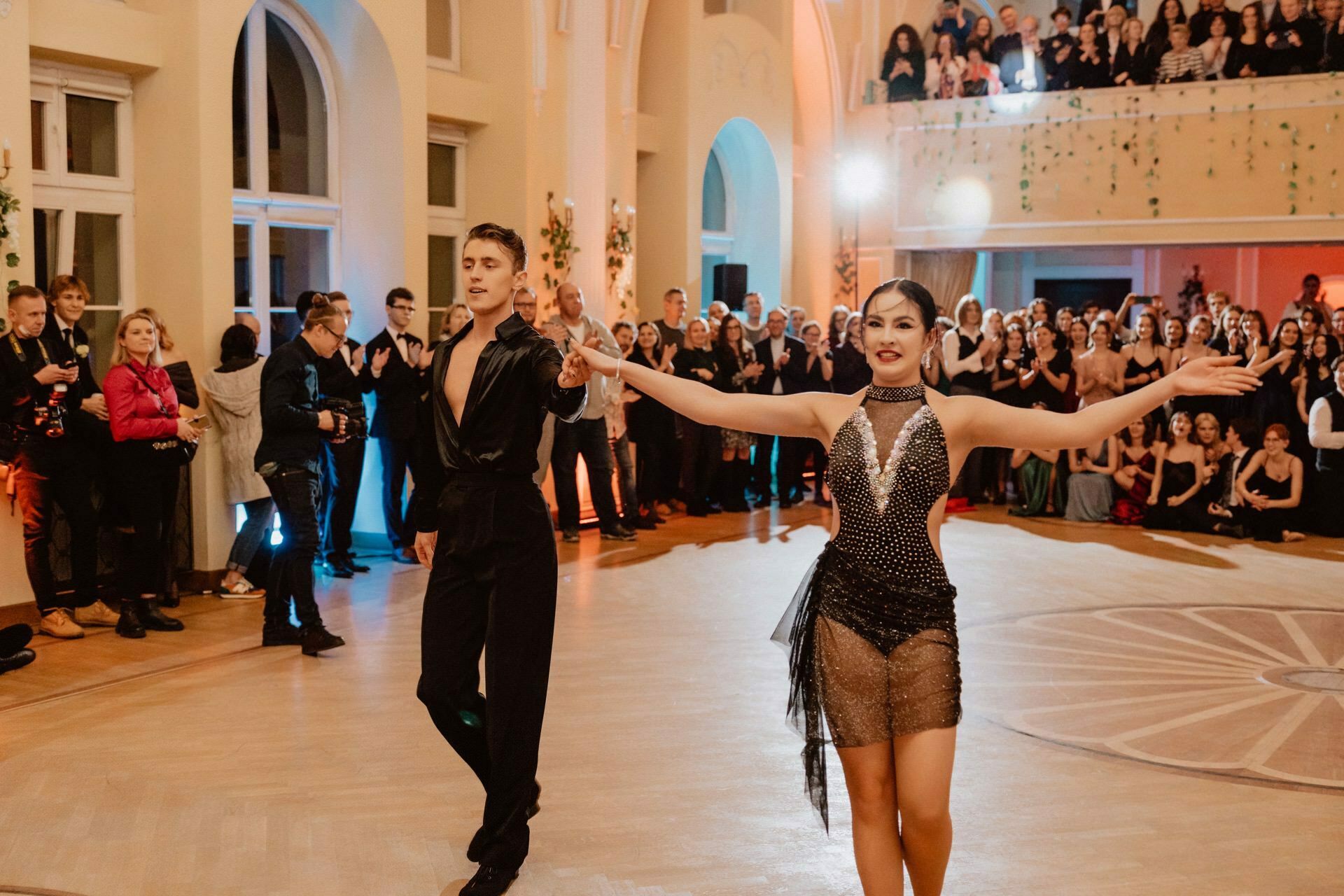 The couple is dancing in a large hall with a crowd of spectators. The man is wearing an all-black outfit and the woman is wearing a glittering dress. She gracefully extends her arms, and the audience behind them claps and watches intently, capturing the essence of this charming reportage of the prom.  