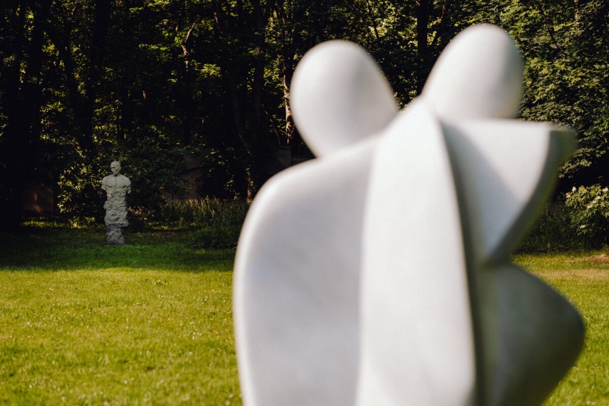 A blurry, modern sculpture of two abstract figures adorns the foreground of a lush green park. In the background, another stone sculpture stands between the trees, spotty rays of sunlight peek through the leaves - beautifully captured by a photographer from Warsaw for a photo essay on the event. 