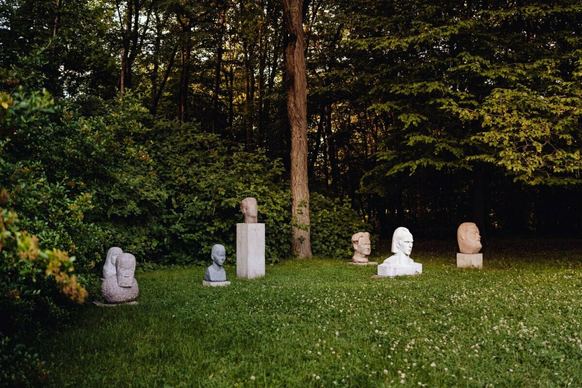 The tranquil outdoor area features a series of stone busts and sculptures arranged in a grassy field. The area, ideal for a photography event, is surrounded by dense green vegetation and tall trees, creating a peaceful, natural backdrop for an art show. 