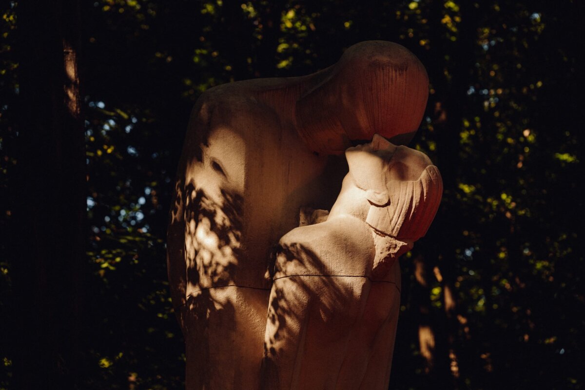 The stone sculpture depicts two abstract human figures embracing and kissing. The figures have smooth, ornament-free surfaces and are bathed in mottled sunlight seeping through the surrounding trees, creating a peaceful and intimate atmosphere reminiscent of moments captured by event photographer Warsaw. 