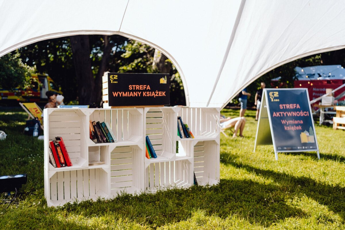 A white bookshelf with several books is located under a tent on the grass. A sign on the bookshelf proclaims "Book Exchange Zone" (Book Exchange Zone), and in the background a second sign reads "Book Exchange Activity Zone." Capture this scene with *photographs at the event* for a perfect*.  