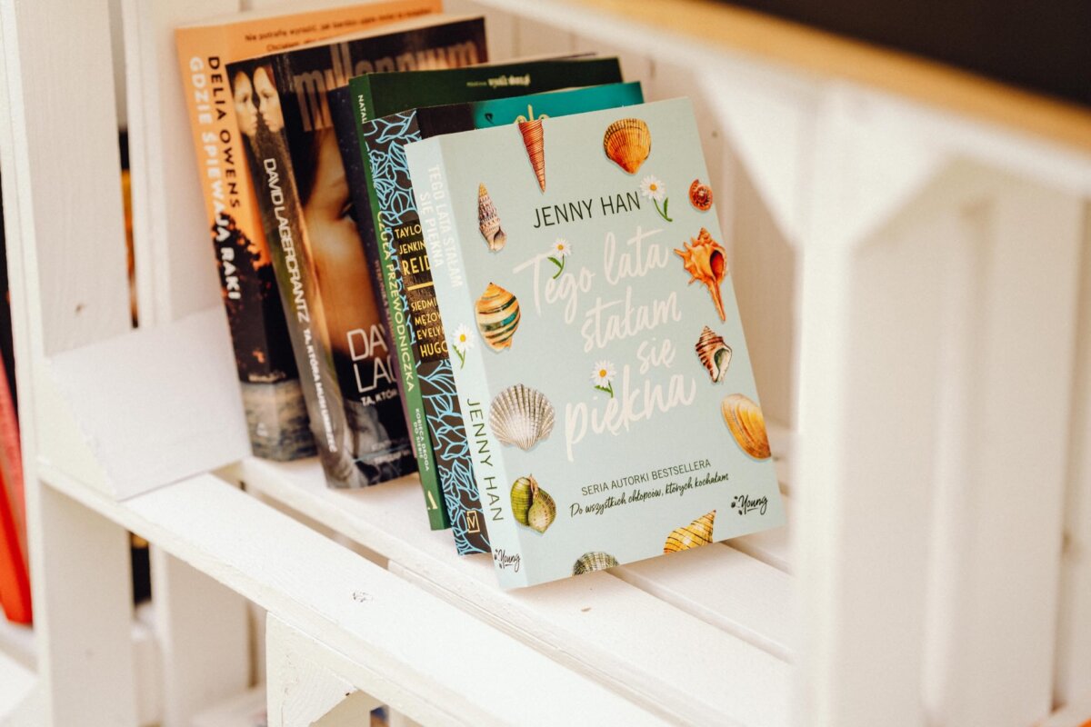 There are several books on the bookshelf, including a prominently displayed book with a shell cover and the Polish title "This Summer I Became Beautiful" by Jenny Han. Other books on the shelf are partially visible with different colored covers, like a photo report of the event captured by an event photographer. 