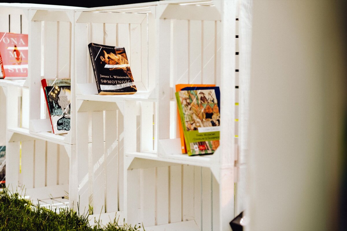 A white wooden bookcase stands on the grass, partially shaded. It's filled with a mix of colorful books, some closed and some open, with different designs and illustrations on the covers. The scene is bright as sunlight filters through, capturing a moment reminiscent of an event photographer Warsaw's snapshot.  