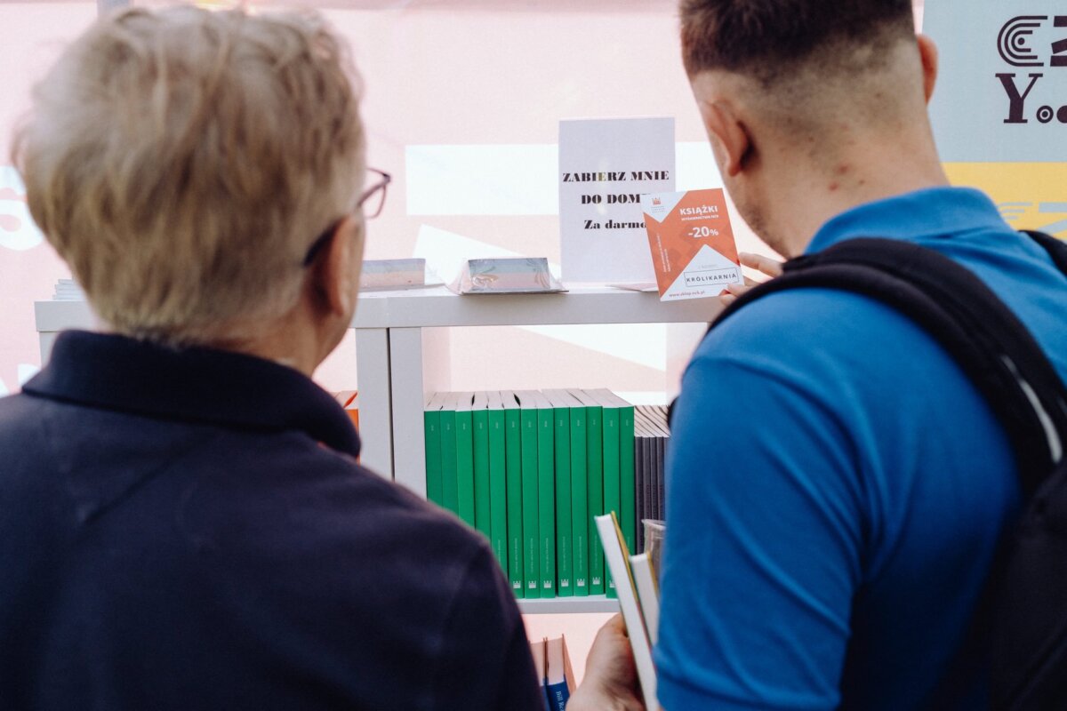 Two people are standing in front of a display with different books. One person has gray hair and glasses, while the other has short hair and a backpack. The exhibit, captured by a photographer from Warsaw, reads in Polish: "Take me home! For free," translated as "Take me home! For free.    