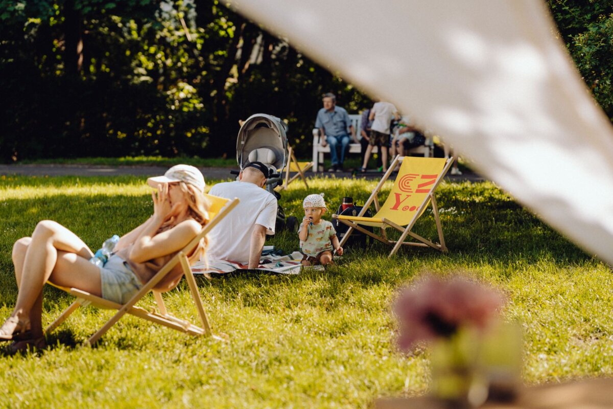 A sunny day in the park with people relaxing on the grass. In the foreground a woman wearing a hat is sitting on a deck chair, perfectly captured by our photographer at the event. Nearby you can see a person with a stroller and a child on a blanket. More people sit in the distance, surrounded by trees and greenery. Delicate flowers provide the setting for this beautiful photo essay of the event.    