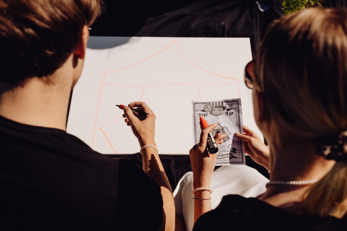 Two people seen from behind collaborate on a drawing. One person holds a piece of paper with a microphone and guitar behind the other person's canvas, which shows the outline of a tent. Both are dressed in dark clothing, and the scene takes place outdoors, perfectly capturing the essence of the event photo essay.  