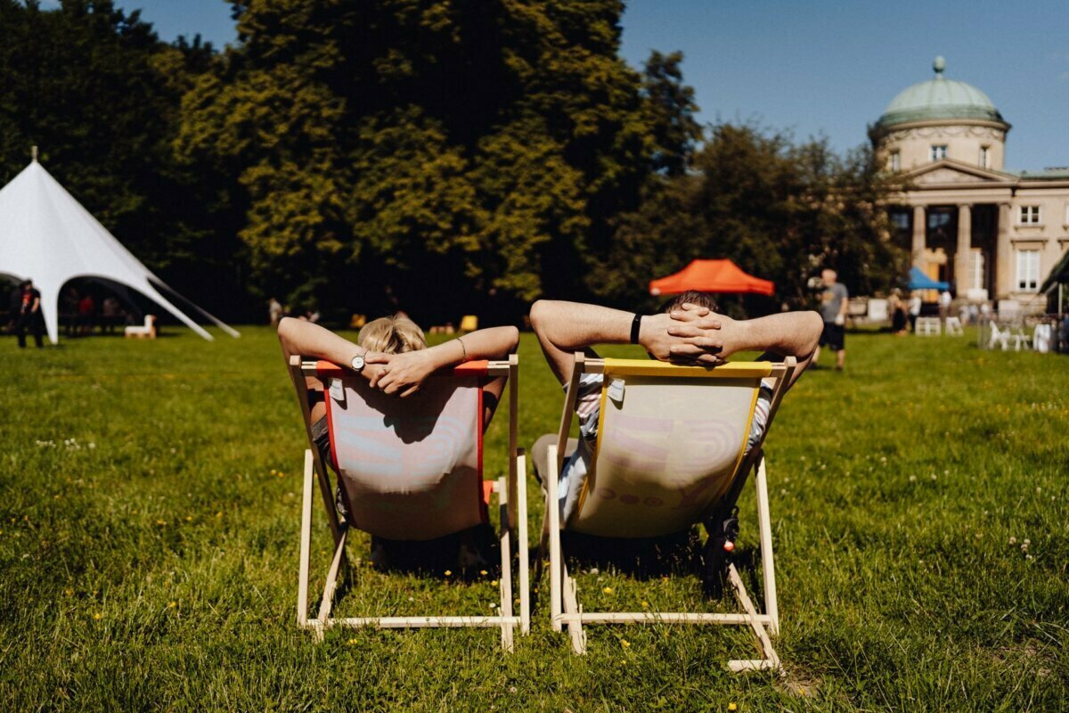 Two people are lounging on colorful deck chairs on a sunny day, with their backs to the camera. They are in an open, grassy field with large trees and a gazebo-like building in the background. A white tent and a few people scattered around announce a vibrant scene, perfect for a photo essay of the event.  