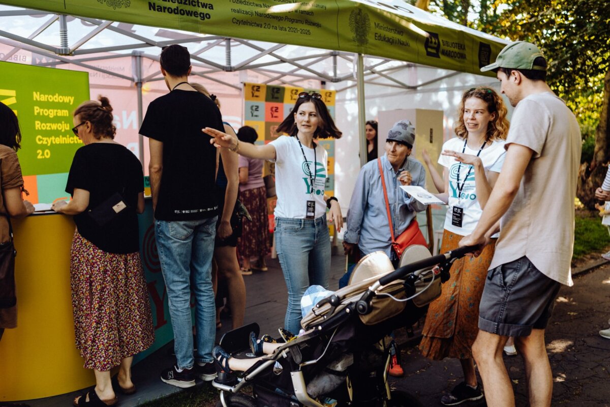 People at an outdoor event booth with a banner reading "National Reading Development Program 2.0." Two women in white T-shirts provide information, others browse through materials. A person pushes a cart, while another stands nearby. The scene is lively and sunny - perfect for any Warsaw-based photographer to capture.   