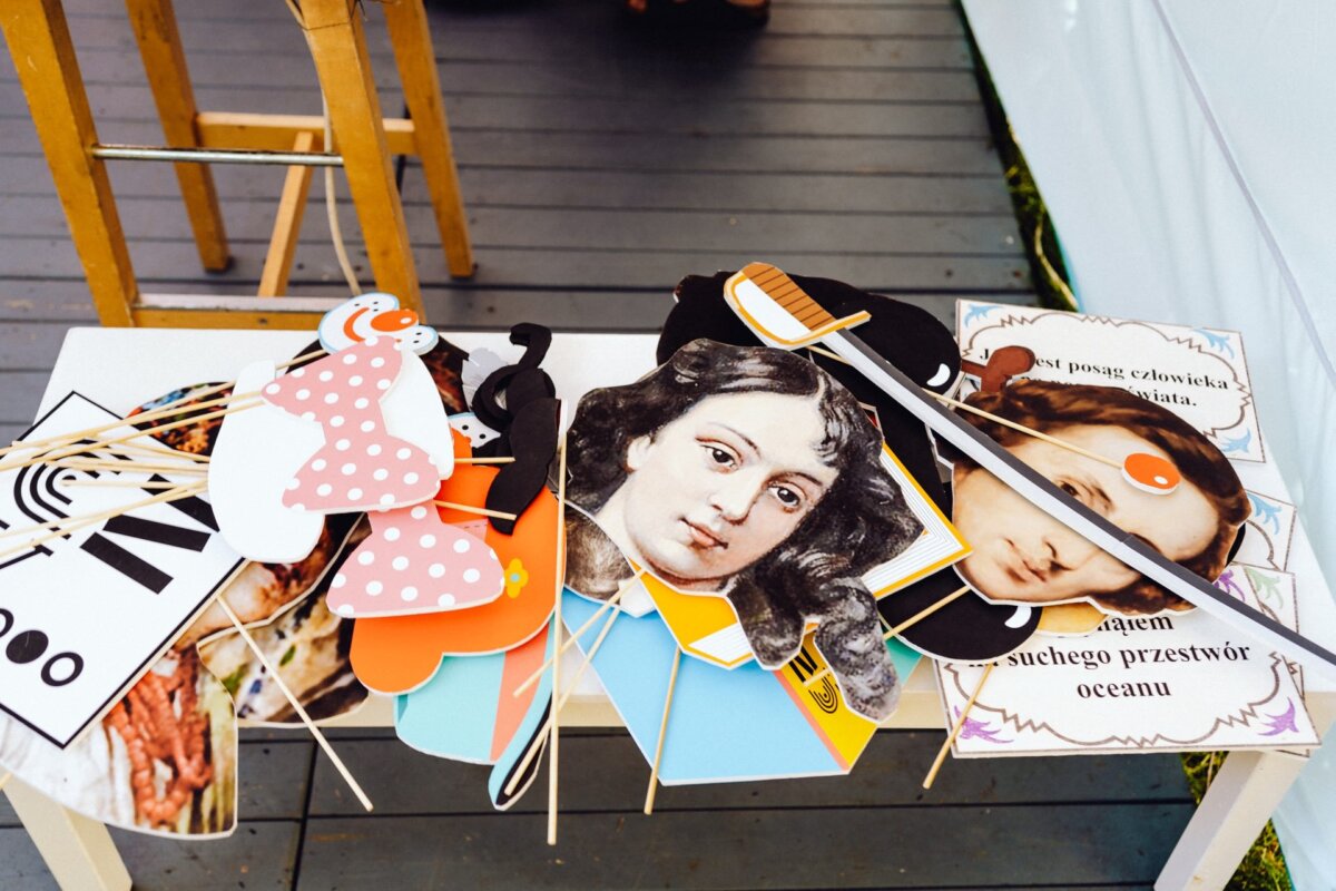 On a white table are colorful paper photo booth props, including cutouts of famous portraits, polka dot bow ties, glasses, a sword and Polish lettering. Nearby, on a wooden terrace, there is a wooden stool - all prepared for a vibrant photo report of the event prepared by a photographer from Warsaw. 