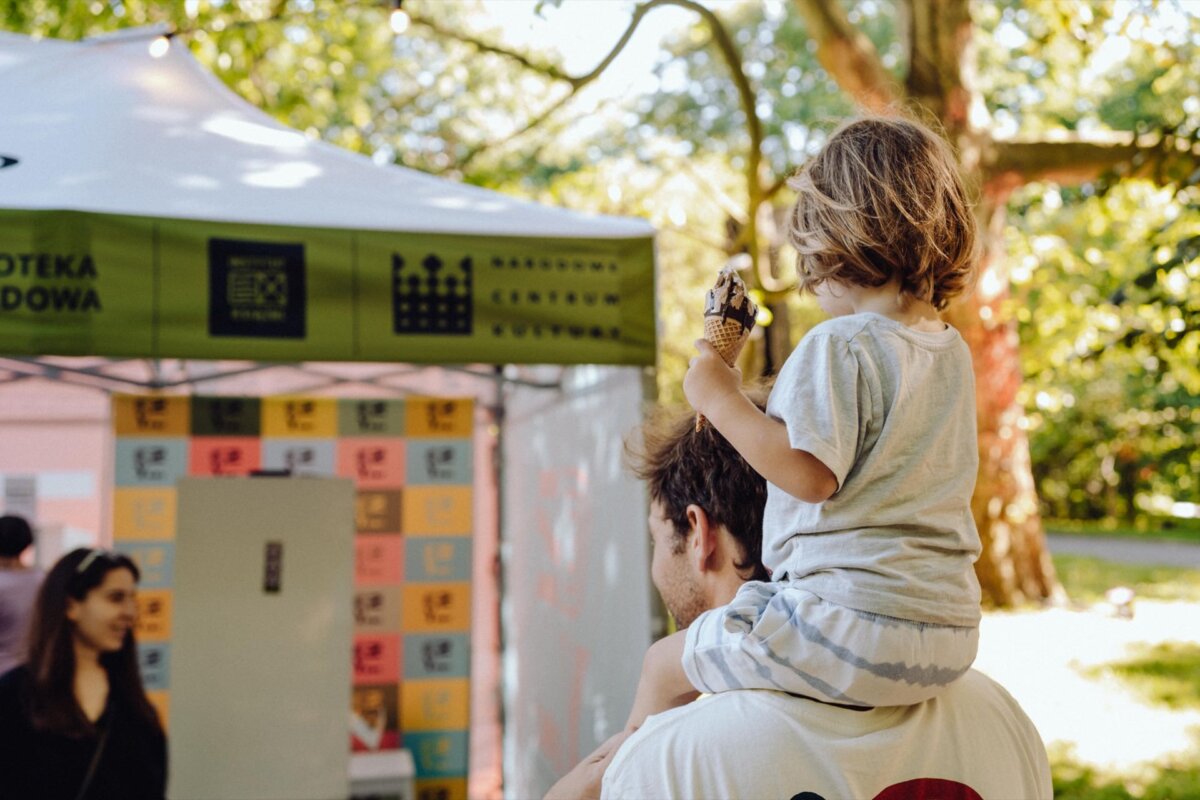 A man carrying a small child on his shoulders stands in front of an outdoor pavilion. The child is holding a chocolate ice cream cone. A tent with a sign reading "NATIONAL LIBRARY" and colorful posters suggests a cultural or library event, beautifully captured by a photographer at an event from Warsaw.  