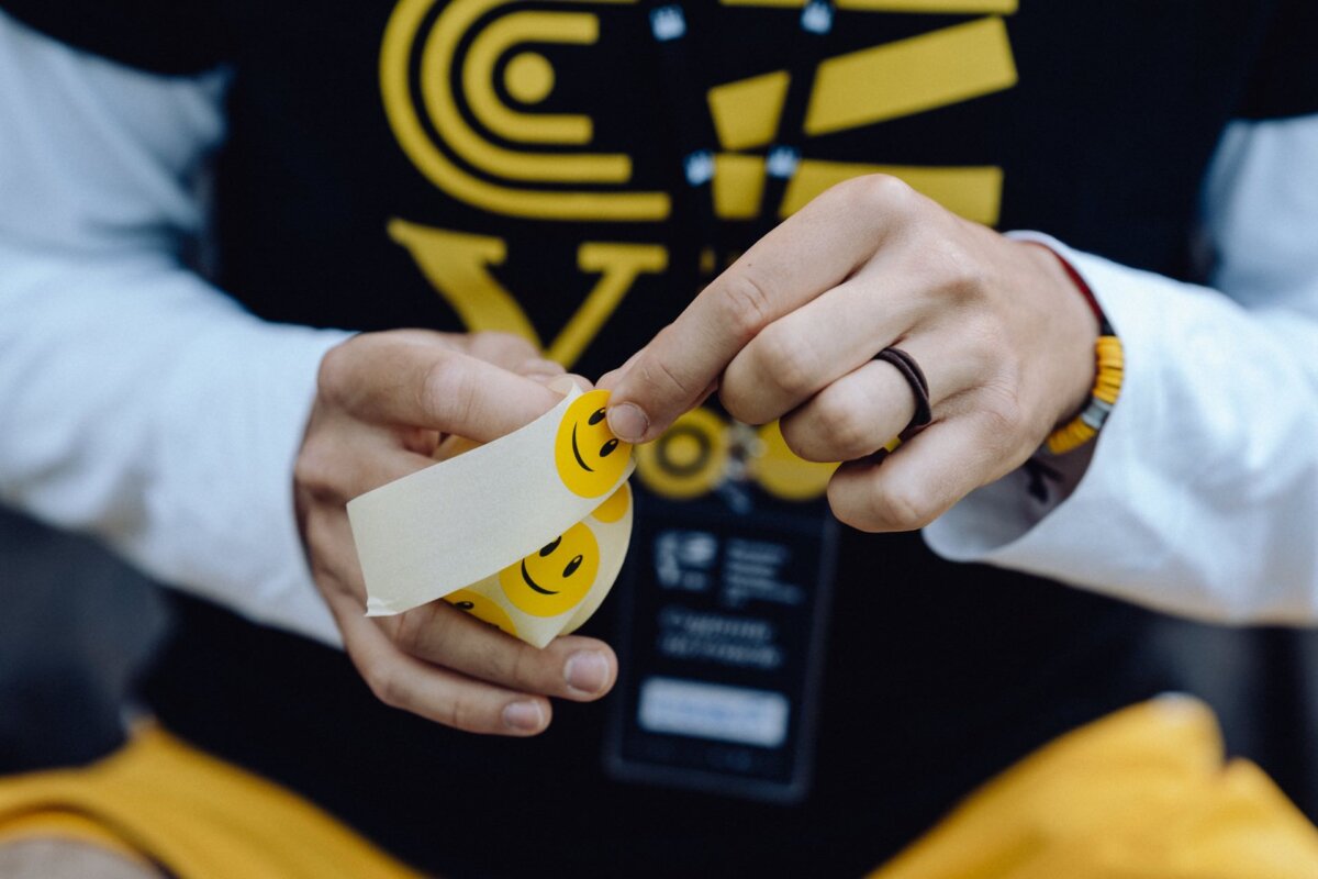 A person dressed in a black and yellow shirt peels off a small yellow sticker with a smiley face from a sheet of identical stickers. With the badge on a lanyard and a ring on his finger, it looks like a photojournalism of the event may have been captured by a Warsaw photographer. 