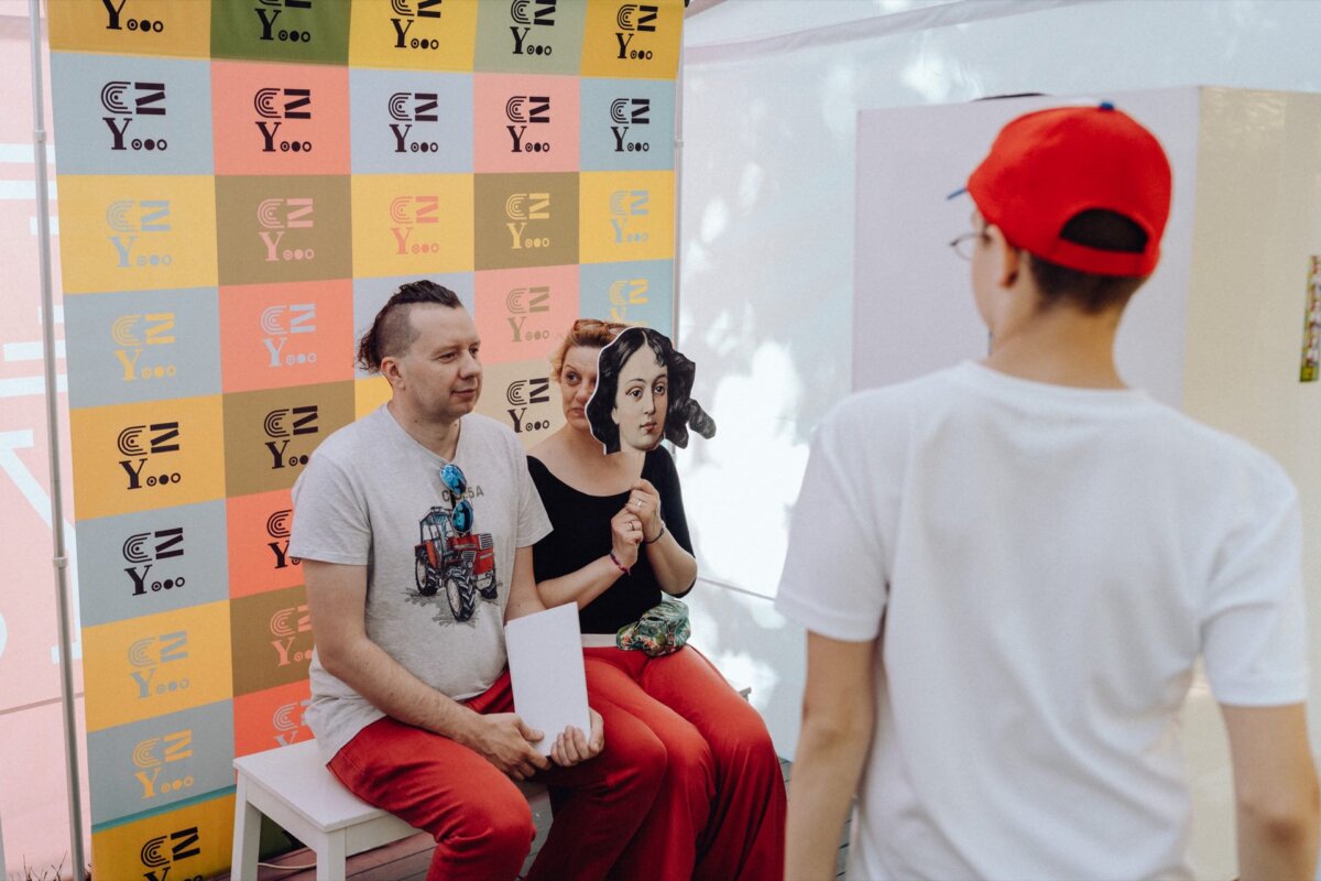 A person in a white shirt and red cap takes a picture of two people sitting on stools against a colorful background with different logos. The seated people are smiling, one person is holding a white piece of paper. It looks like everyone is at some kind of event or gathering, captured by the event photographer Warsaw.  