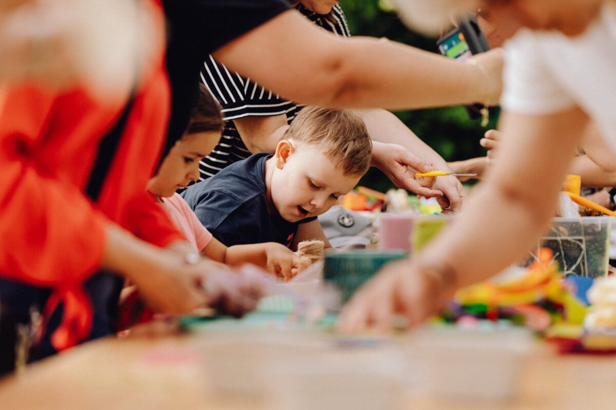 Children and adults take part in group craft activities at a table filled with art supplies. The children seem to be focused on their projects, while the adults help and engage with them. A variety of colorful materials can be seen in the workspace, creating a perfect photo-op of the event.  