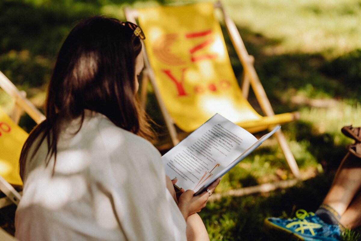 A person with long hair sits on the grass and reads a book. Yellow deck chairs are visible in the background, casting shadows on the floor. The scene, perhaps captured by an event photographer Warsaw, brings to mind a calm, sunny day in a park or garden.  