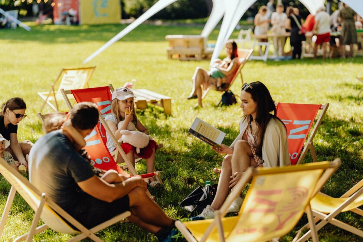 People relaxing on a sunny day, sitting on colorful deck chairs on a grassy area. One person is reading a book, while others are loosely chatting. In the background, more groups sitting around wooden tables under a large white canopy, beautifully captured by event photographer Warsaw. The stage is surrounded by trees and greenery.   