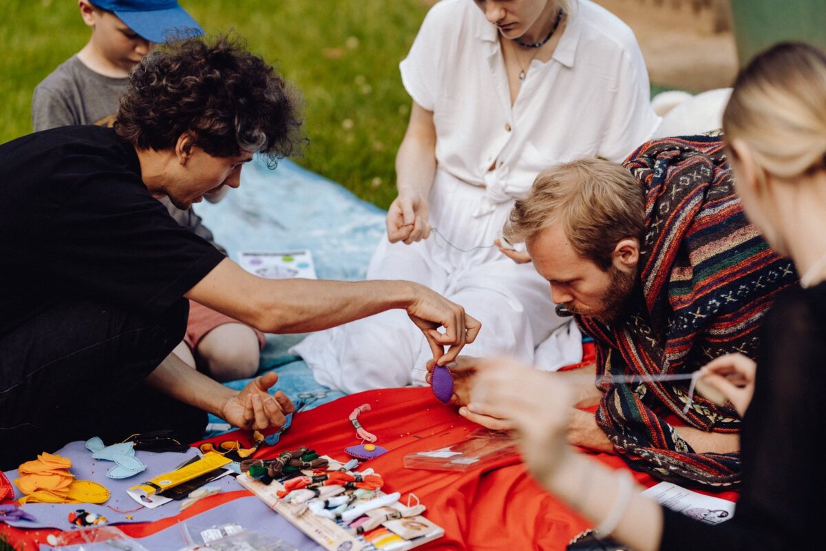 A group of people sit outside on a blanket and do crafts. One person passes a small purple object to another, and various handicraft utensils, including thread, scissors and cloth, are scattered around. Trees and grass are visible in the background. Perfect for a photography party!   