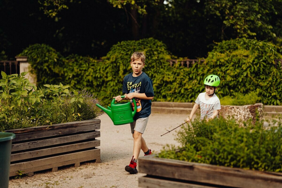 A boy carrying a green watering can and a spray bottle walks along a path in a garden with raised beds. A younger child in a green bicycle helmet, holding a stick, follows beside him. Surrounded by greenery on this bright sunny day, it is the perfect scene to capture any *event photographer Warsaw*.  