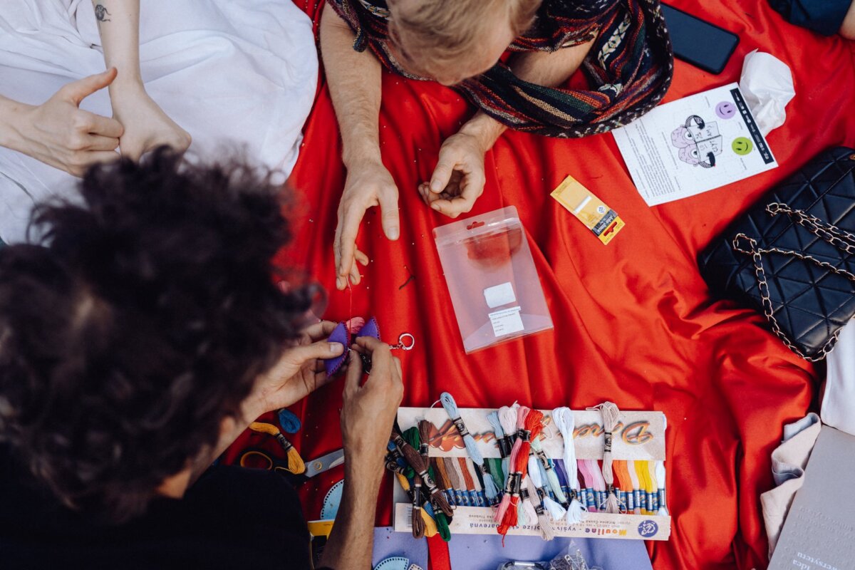 People sit on a red blanket and engage in handicrafts. Various colored yarns, a clear plastic ziplock bag, scissors and craft tools are spread out. A black bag and an instruction booklet can also be seen on the blanket - a perfect scene for a photo essay of the event by a photographer from Warsaw.  