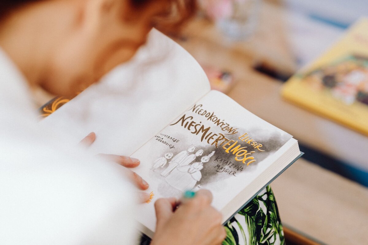 A person with blurred facial features is holding a Polish book entitled. "Uncommon Lessons in Germanness" by Katarzyna Kęcik. The book's cover features grayscale illustrations of people. The blurred background shows colorful objects and another book, resembling a photo report of the event.   