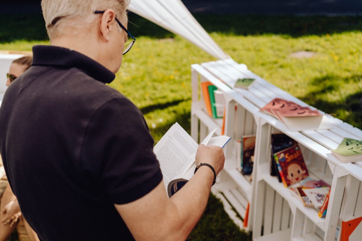 A person with short, gray hair and glasses is reading a book outside next to a white box-shaped shelf filled with more books. The scene takes place on a grassy lawn, illuminated by sunlight, which was beautifully captured by a photographer from Warsaw. 