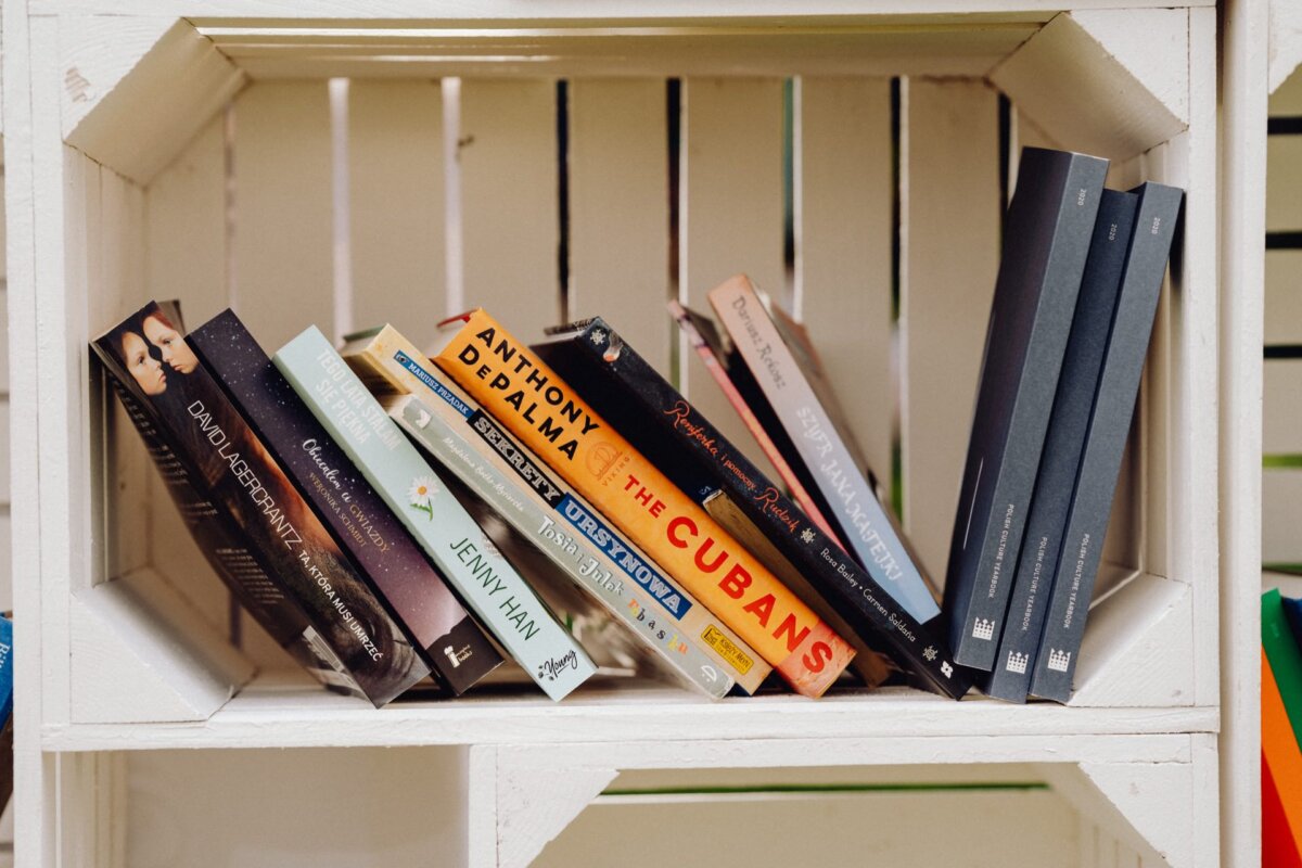 The wooden shelf will hold a variety of books arranged both vertically and horizontally. Some visible titles include "The Cubans" by Anthony DePalma, "P.S. Still Love You" by Jenny Han and "Out of Darkness" by Ashley Hope Pérez. The bookshelf, reminiscent of a photo event, has a rustic, whitewashed look.  