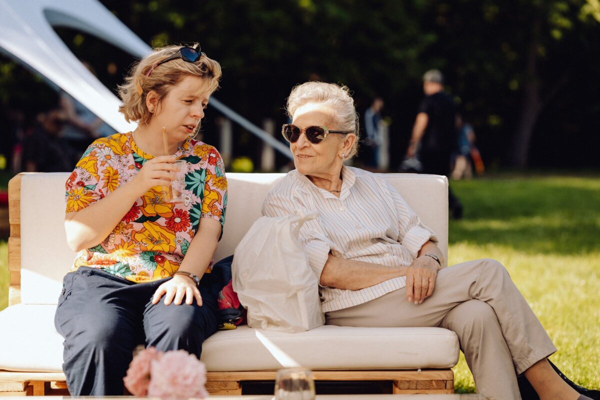 Two women are sitting on a sofa outside having a conversation. The younger woman, wearing a colorful floral shirt and holding a drink, faces an older woman wearing a striped shirt and dark sunglasses. Captured by a photographer from Warsaw during an eventful day in a sunny park with lush green grass in the background.  