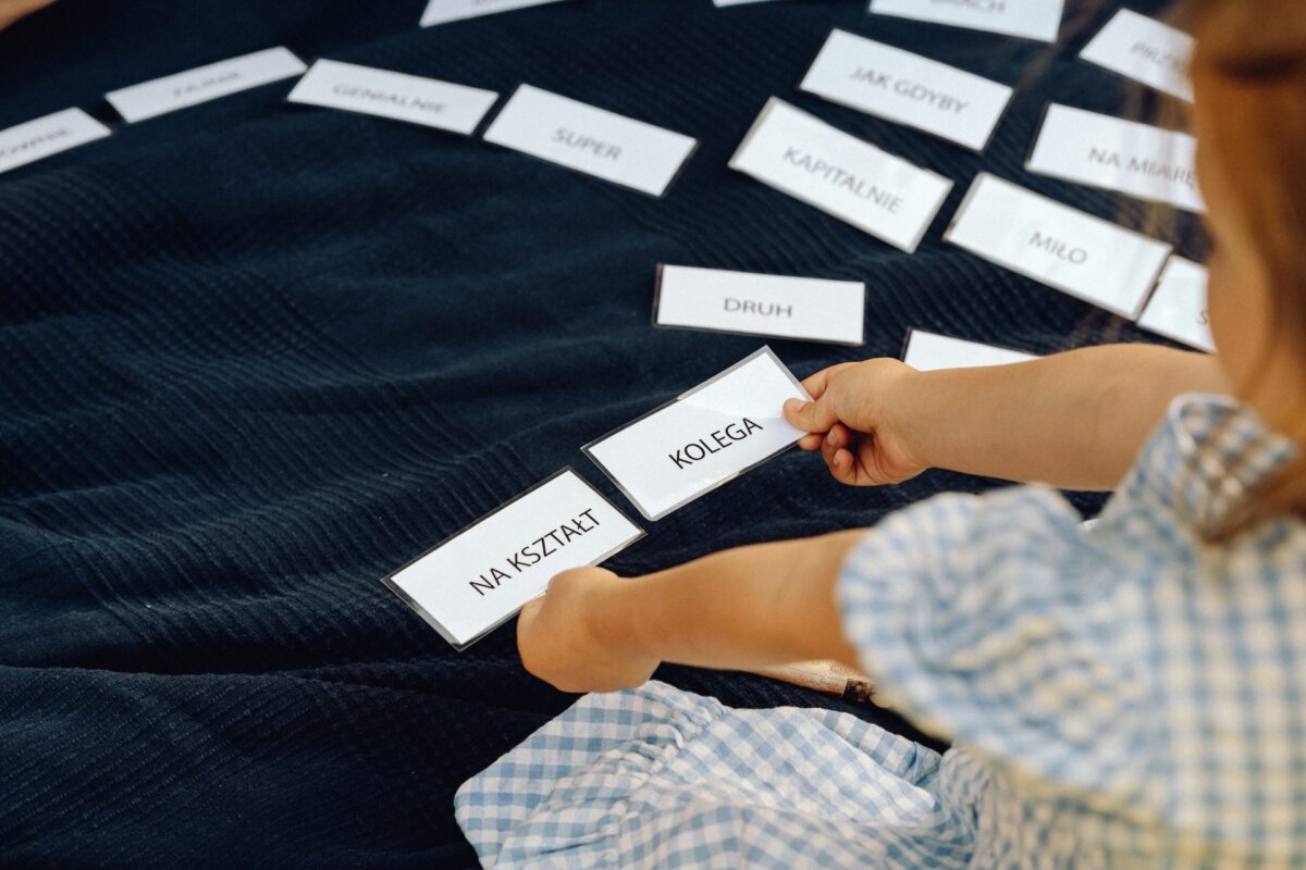 A child in a checkered dress arranges cards with Polish words, including "COLLEGE" and "FOR SHAPE," on a dark cloth. More marked cards are scattered around, indicating a learning or matching activity. This scene, captured by a photographer from Warsaw, beautifully shows the joy of discovery.   