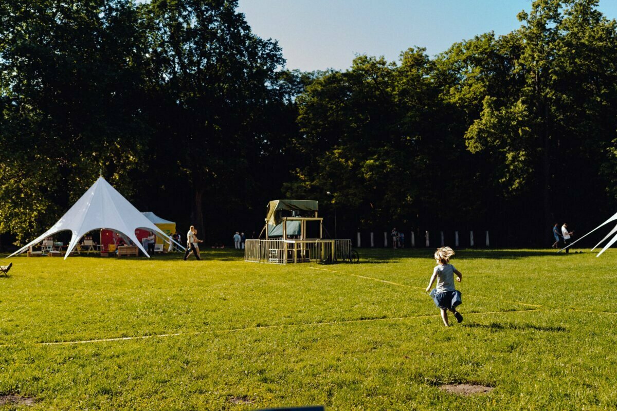 A young child runs across a grassy field with a playground and a large white tent in the background. Near the tent and among the trees, several people can be seen enjoying the sunny day. The scene is set in a park with lush green trees and grass, perfect for a photo essay of the event by a photographer from Warsaw.  