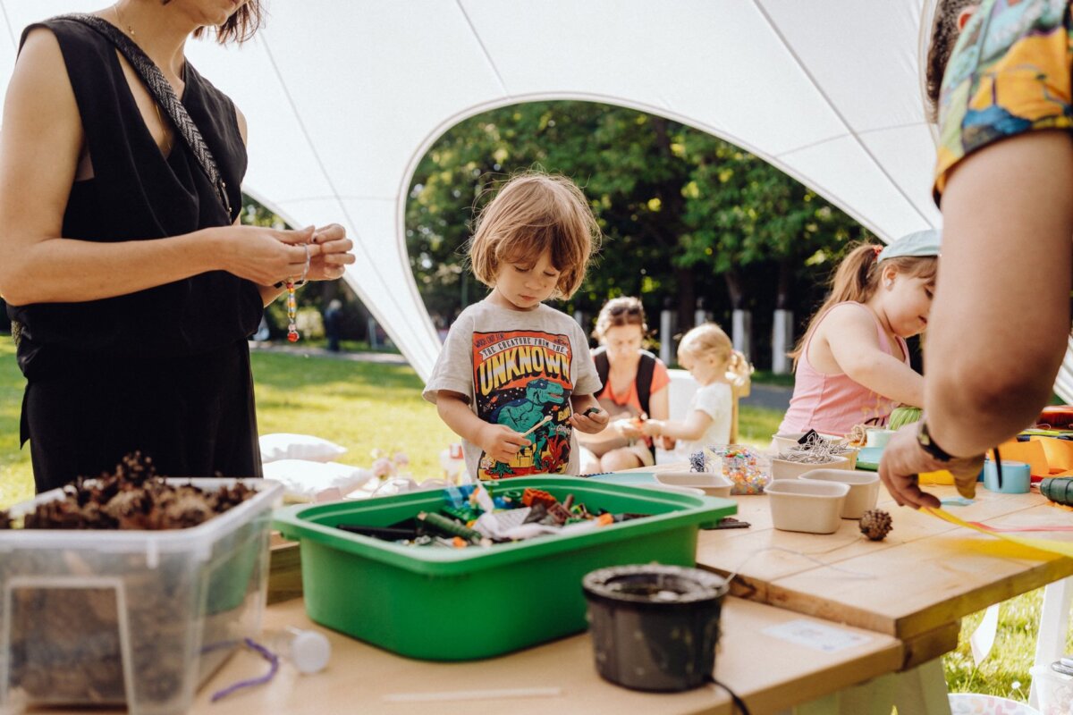 On a sunny day, a group of children and adults are doing craft activities under a tent. One child in the middle, wearing a colorful T-shirt, focuses on a craft. Tables are filled with utensils, including a green tub and various art materials. Trees are visible in the background, beautifully captured by the photographer for the event.   