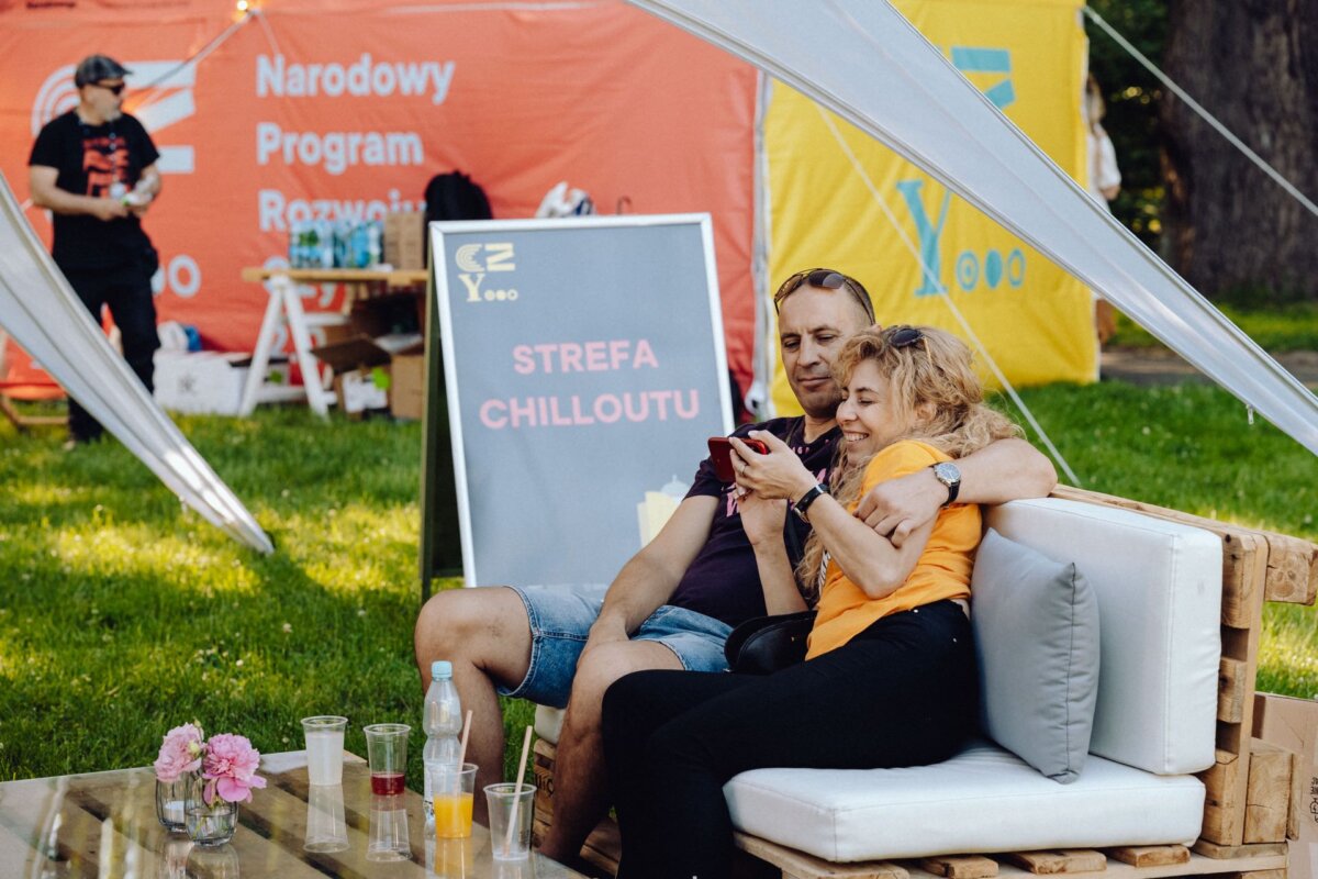 A man and a woman sit together on a padded bench outside, the man puts his arm around the woman's shoulder as she looks at her phone. They are in a quiet, shaded area, with a sign behind them reading "Chillout Zone." On a table in front are flowers, water bottles and drinks - perfectly captured by a photographer from Warsaw.  