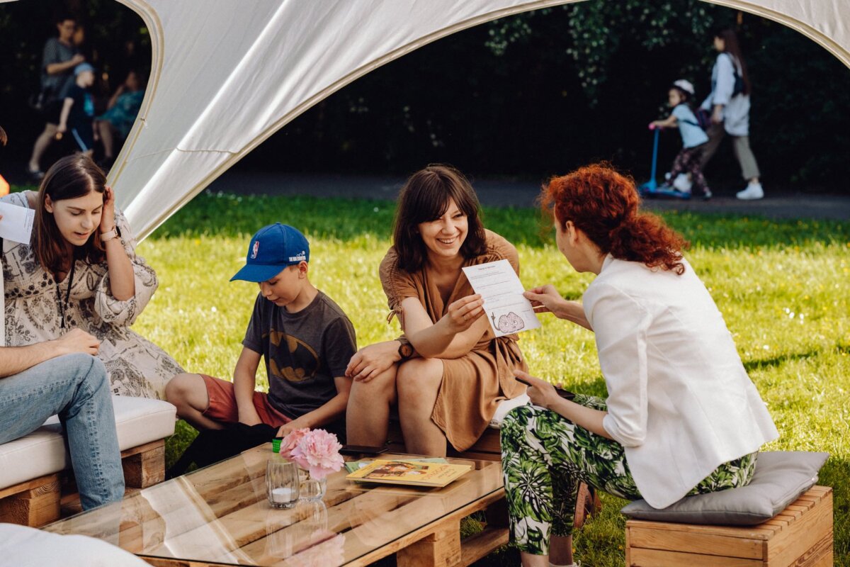 A group of people are sitting outside under a canopy. A woman in a beige dress is showing a piece of paper to another woman in a white jacket and patterned pants. A child in a blue cap and Batman T-shirt sits next to them, and another woman sits next to them, all beautifully captured in a photo report of the event by a photographer from Warsaw.  