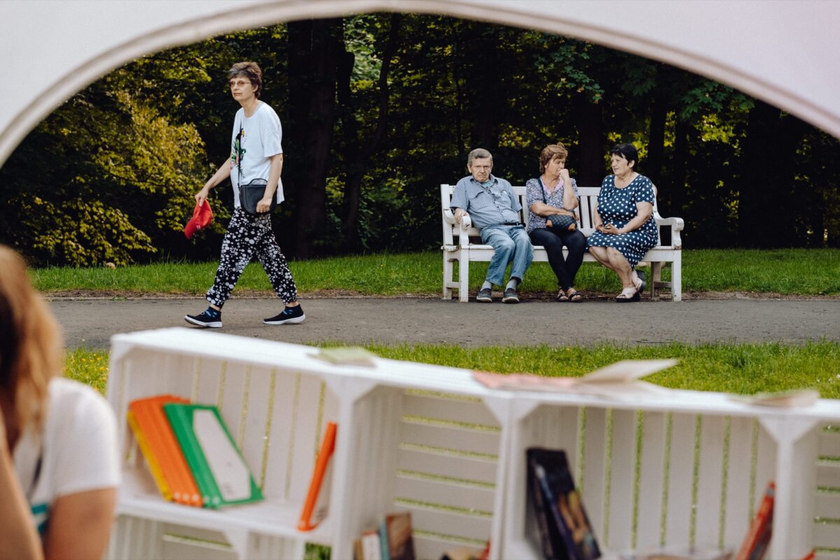 The Warsaw-based photographer captured a person dressed in black-and-white patterned pants and a white shirt walking past three people sitting on a park bench. In the foreground, open shelves display colorful books, while trees and grass form the background. 