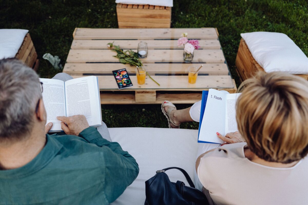 A man and a woman are sitting on seats outside and reading books. Between them, on a low wooden table, stand two glasses of orange juice with straws, a jar with a candle, a bouquet of flowers and an electronic device. In the background you can see grass and cushions. It's a perfect scene for a photo report of the event by a photographer from Warsaw.   