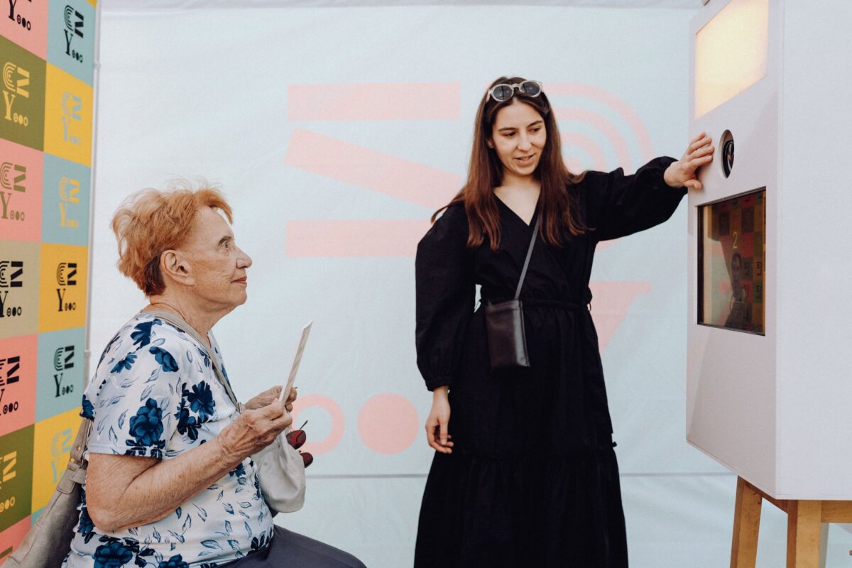 An older woman with short, reddish hair, sitting and holding a piece of paper, looks at a standing woman with long brown hair operating a booth or machine. The younger woman, probably a photographer from Warsaw, is wearing a black dress and shoulder bag while interacting with the machine. 