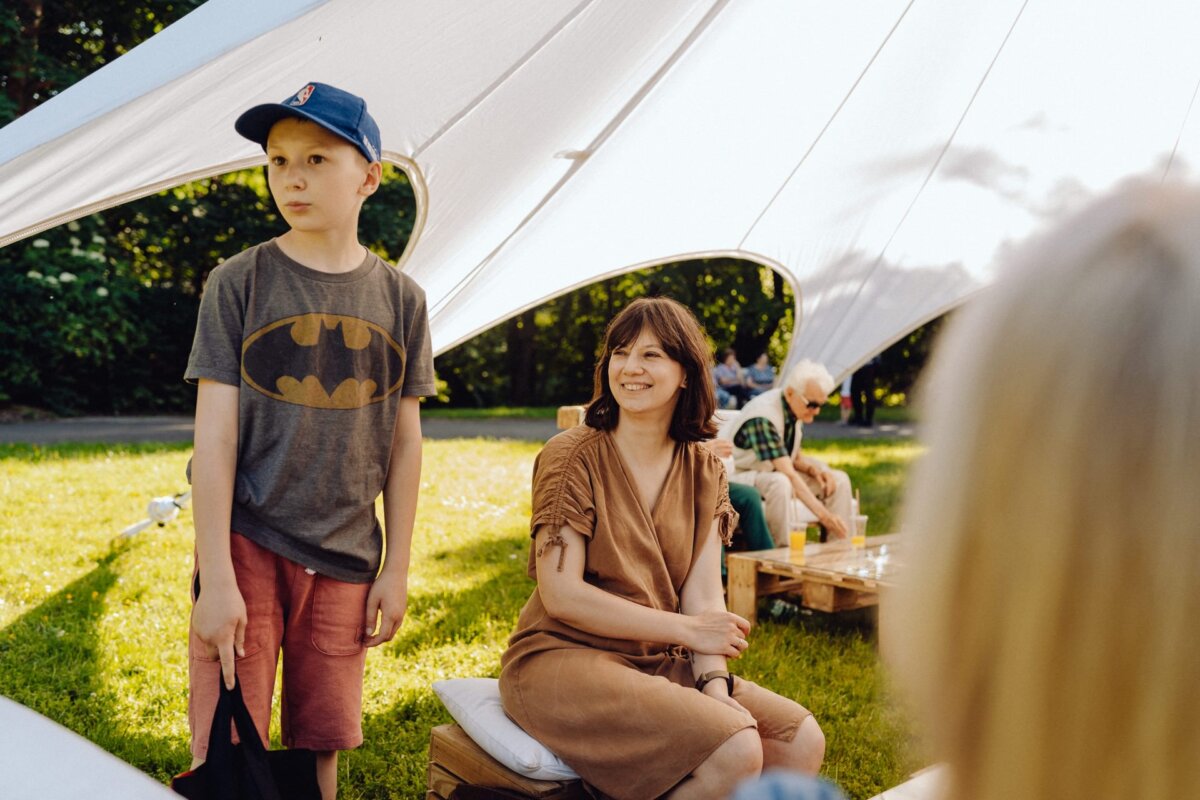 A boy dressed in a Batman T-shirt, red shorts and a blue cap stands next to a woman in a brown dress, who is sitting on a cushion under a white canopy. Captured by a talented photographer from Warsaw, the scene takes place outdoors on a sunny day, against a backdrop of trees and other people. 