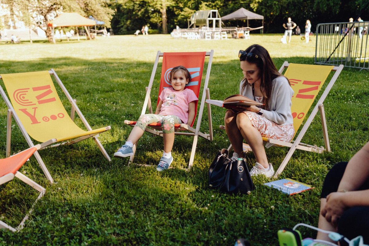 A woman is reading a book while sitting on a folding chair on the grass. Next to her on another folding chair sits a smiling child, dressed in a pink shirt and patterned pants. In the background of the Warsaw event, several empty folding chairs and people can be seen, all in the park.  