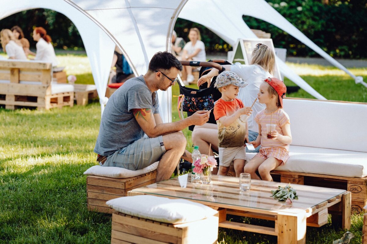 A man sits on an outdoor couch with his two children and shares snacks and drinks on a sunny day. They are under a white canopy, while others are relaxing in the background on similar wooden furniture. The scenery is reminiscent of a simple picnic or outdoor gathering captured by a photographer at an event.  