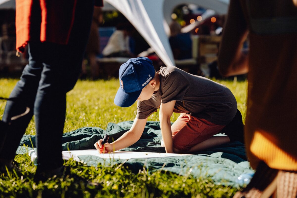 A young boy wearing a blue cap, gray shirt and red shorts kneels on a blanket on the grass and draws with a marker on a large sheet of paper. Sunlight filters through the trees, creating a warm and inviting atmosphere. Captured by a photographer from Warsaw, other people and a white canopy can be seen in the background.  